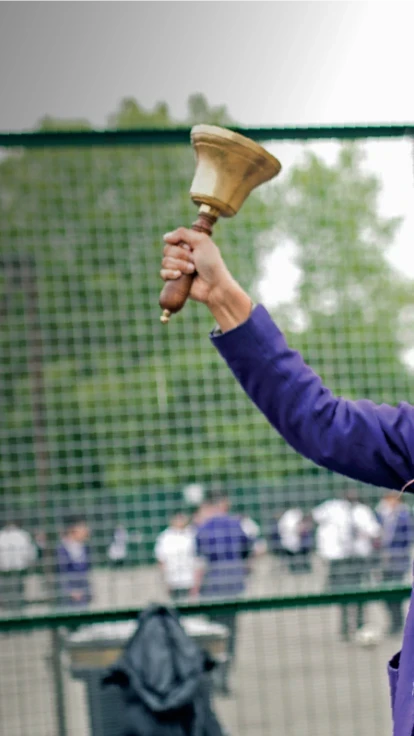 Smiling schoolgirl in a purple uniform ringing a handbell in a school playground, symbolising achievement and celebration.