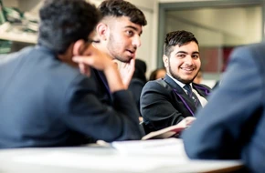 Three secondary school boys in uniform engaged in a classroom discussion, smiling and listening attentively.