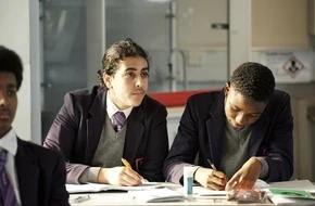 Two secondary school students in uniform working at their desks in a classroom.