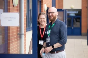 Two Teach First delivery partners standing outside a school building, smiling and wearing ID lanyards.