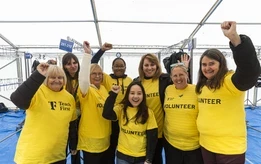 A group of smiling volunteers wearing yellow "Teach First Volunteer" T-shirts raise their arms in celebration at the 2024 Run the River event.