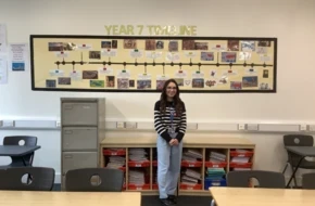 Thea, a history teacher, stands in her classroom in front of a Year 7 timeline display, smiling at the camera.